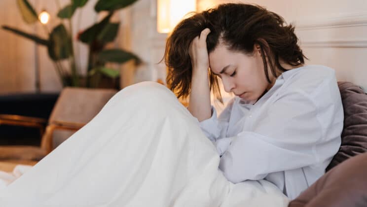 Depressed woman touching hair and closing eyes while resting on bed under warm blanket at home